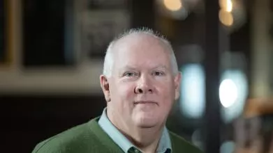 Professor Timothy Power has short white hair and is wearing a green jumper and light blue shirt