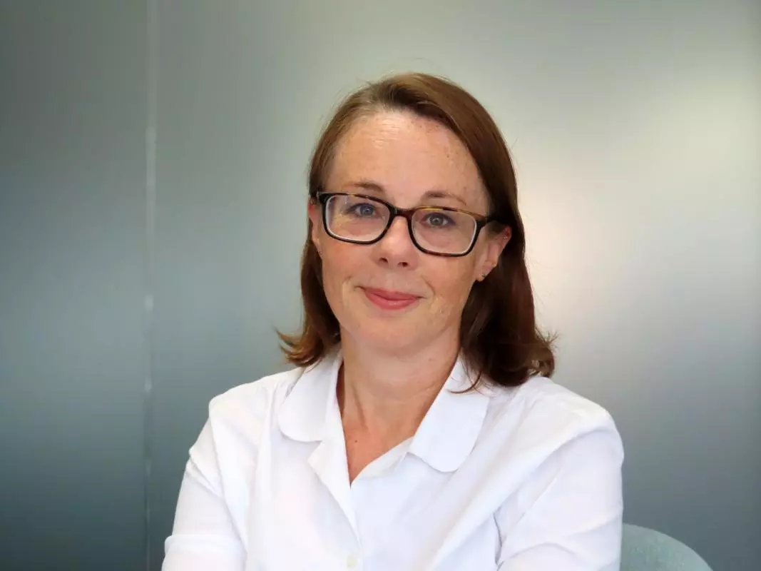 Rachel has shoulder-length brown hair and dark glasses. She is wearing a white shirt.