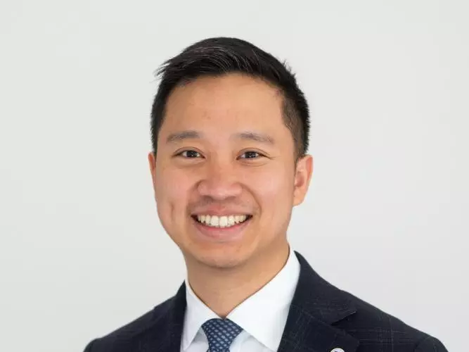 Dr Juin has short black hair. He is wearing a white shirt, blue tie and navy suit jacket. 