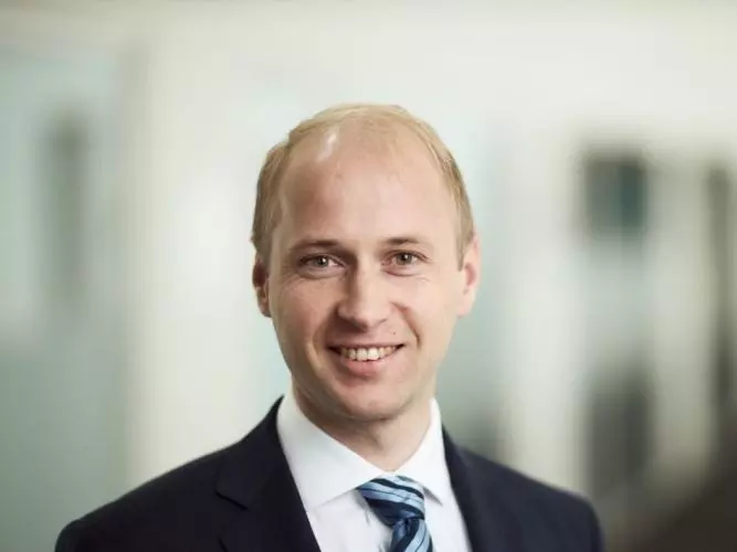 Dr Knesl has fair hair and light brown eyes, and is smiling. He is standing in a corridor, wearing a navy suit, white shirt and blue striped tie. 