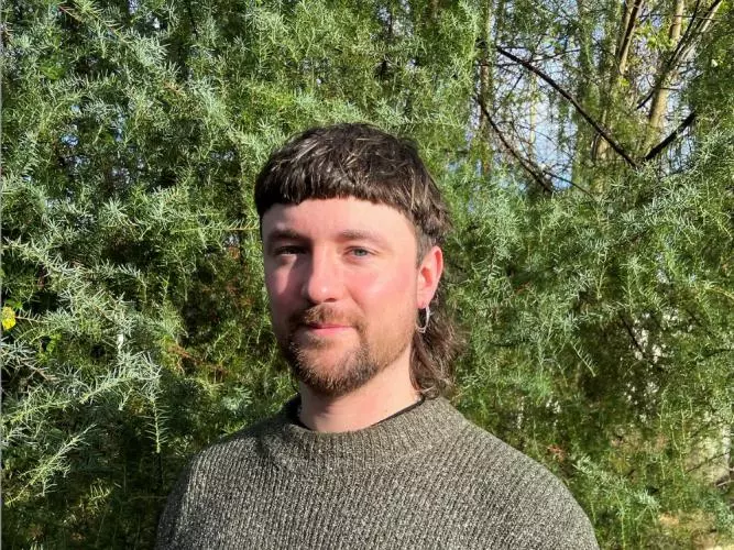 Dr Turnbull has brown hair styled in a mullet. He is wearing a green knitted jumper and standing in front of some foliage. 