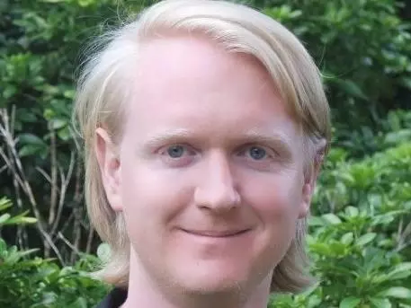 Dr Wainwright has long blonde hair and is wearing a black t-shirt and blazer. He is standing in front of foliage and smiling. 