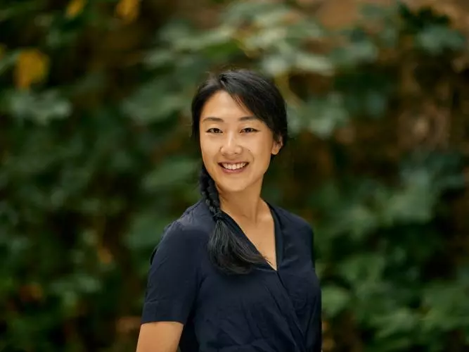 Dr Shi has long dark hair tied into a plait, falling over her shoulder. She is wearing a navy blouse and standing in a garden setting.