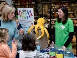 Children and adult at welcome desk to the Festival of Social Science, talking to event lead. Table has maps and leaflets on, and a feedback box with a yellow toy octopus on top. 