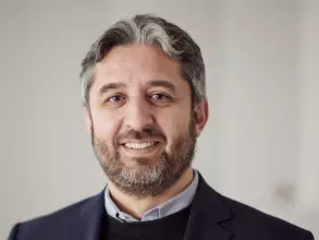 Dr Mahdi has short grey and dark hair and a short beard. He is wearing a shirt, navy jumper and blazer. 