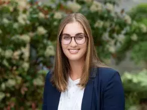 Dr Danica Sims is standing in front of a bush with white flowers. She has straight brown hair and glasses. She is wearing a white blouse and navy blazer. 