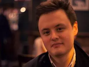 Dr Howell has short brown hair. He is sat inside, wearing a white shirt and dark jumper under a dark coat. 