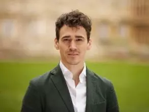 Dr Stephany is standing in front of a large patch of grass. He has short brown hair and is wearing a white shirt and grey blazer. 