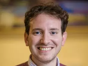 Dr Rampazzo has short brown curly hair. He is wearing a dark pink jumper and smiling widely at the camera.