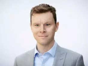 Professor Greg Taylor has short brown hair. He is wearing a light blue shirt and grey blazer. 