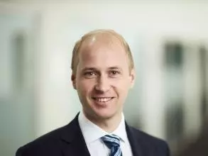 Dr Knesl has fair hair and light brown eyes, and is smiling. He is standing in a corridor, wearing a navy suit, white shirt and blue striped tie. 