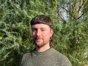 Dr Turnbull has brown hair styled in a mullet. He is wearing a green knitted jumper and standing in front of some foliage. 