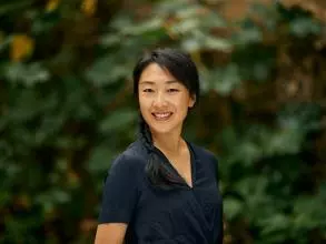 Dr Shi has long dark hair tied into a plait, falling over her shoulder. She is wearing a navy blouse and standing in a garden setting.