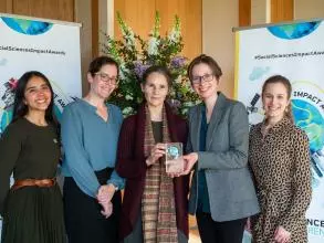 The OPHI team smile and hold their prize trophy. In the background is the Impact Awards branding and a flower arrangement.