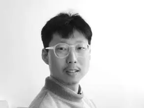 Dr Park has short black hair and is wearing light, round-rimmed glasses and a turtleneck jumper. He is sat at a desk and turned slightly to look at the camera.