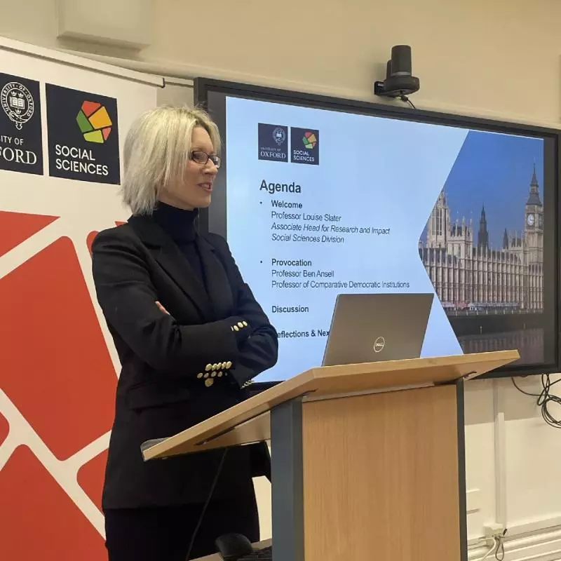 Professor Louise Slate stands at a lecture podium in front of Social Sciences Divisional branding and a slide of the agenda for the Democracy seminar