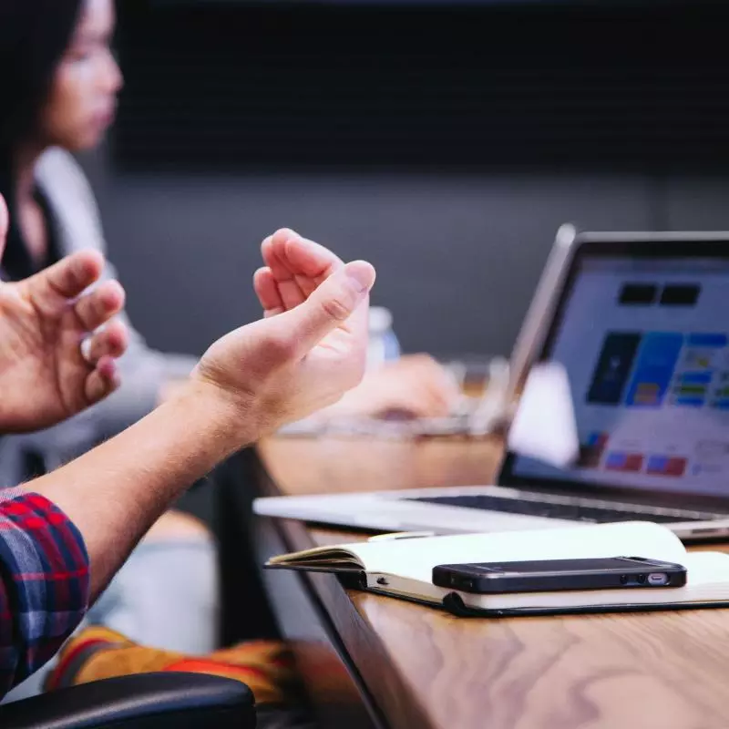 A person sitting in front of their laptop and an open book gestures as if in discussion with their co-workers who are in the background