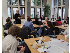 A male academic addresses a room full of researchers who listening to him attentively, and seated around tables that are covered in pens and paper, brainstorming activities, and lunch