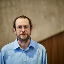 Man with long hair in a ponytail and beard, glasses and blue shirt stands in front of camera