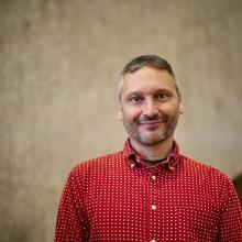 Man with black and grey hair and stubble and red shirt looks to the camera