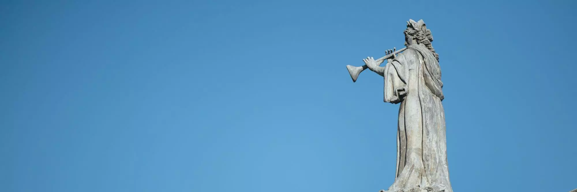 A statue of a robed figure blowing a trumpet, positioned on the roof of a building against a bright blue sky