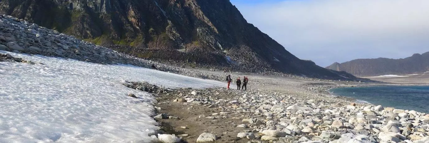 Three people walk across a beach in the artic. There is snow on the left and a moutain in the background.