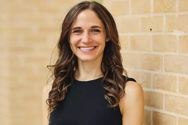 Dr Hawbaker has long wavy brown hair is wearing a black sleeveless top, and is smiling whilst standing by a brick wall.